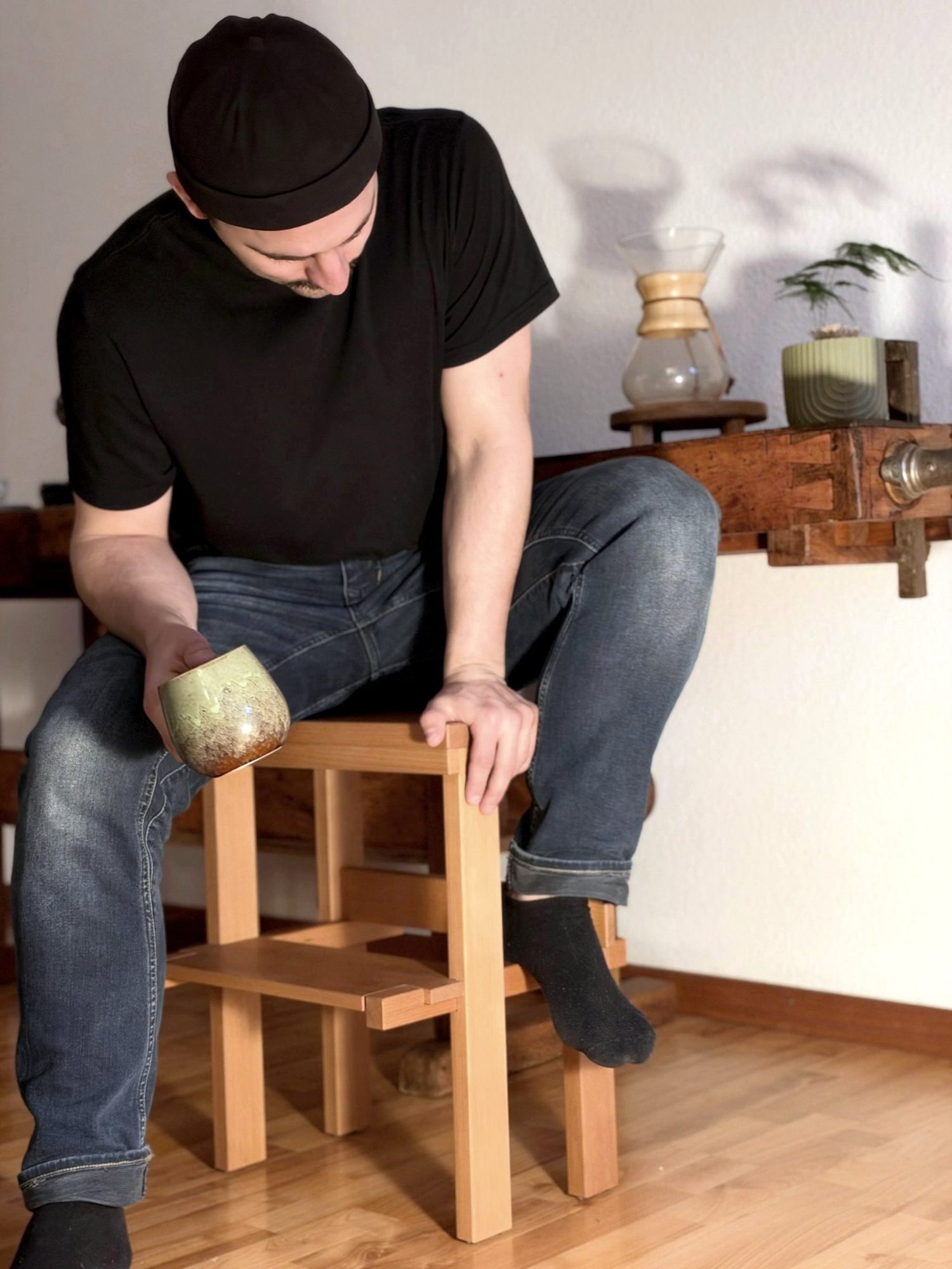 Eugen sitzt auf dem fertigen Hocker vor seiner Hobelbank, mit einer Tasse in der Hand