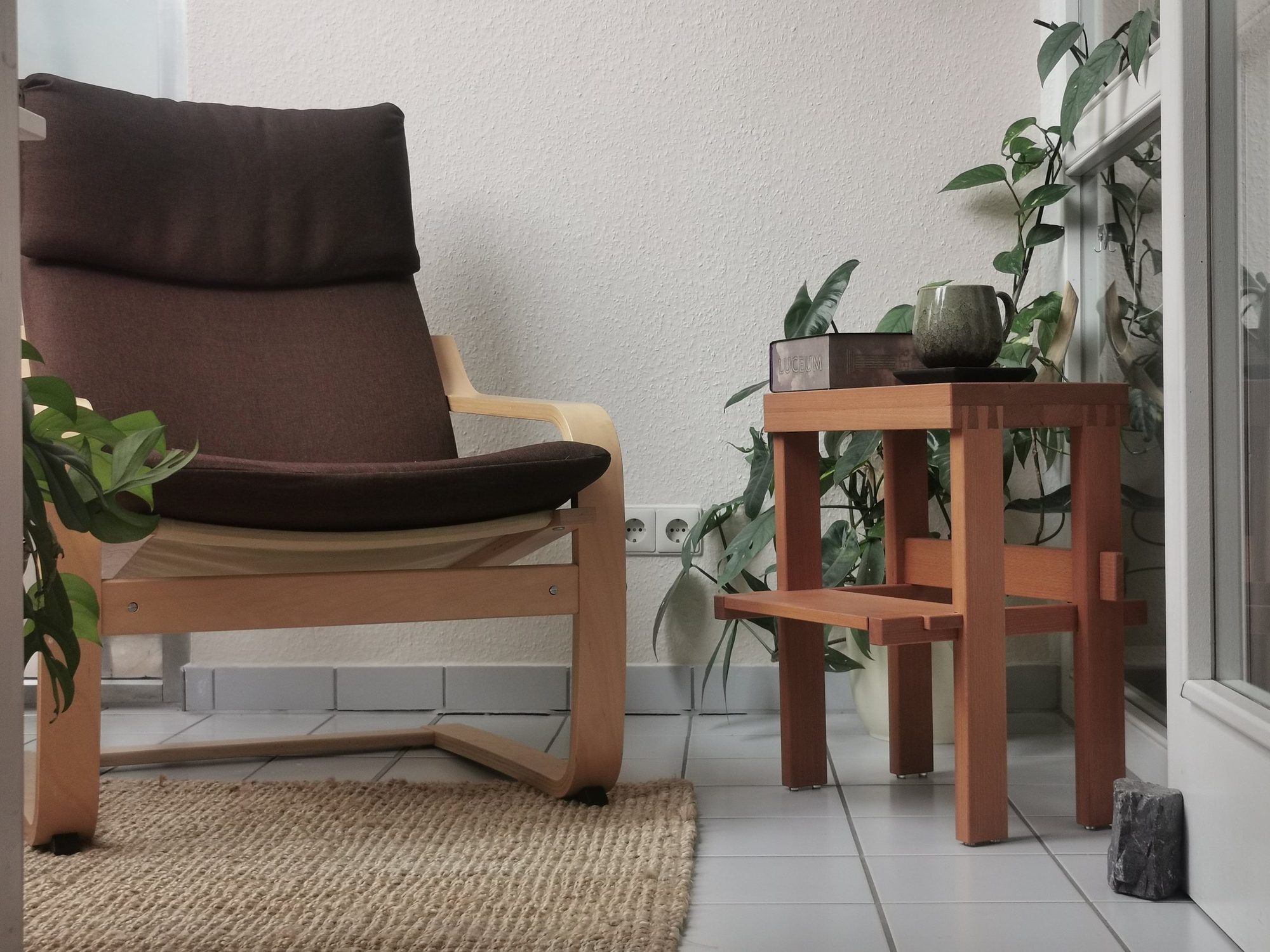 Der Hocker aus gedämpfter Buche als Beistelltisch neben einem Sessel, darauf eine Tasse und ein Buch